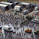 A high-definition photograph depicting an evacuation scene in the Russian border area. Visible signs of heightened security are evident, with miscellaneous structures, roadblocks and emergency services vehicles. Civilian characters represent diverse genders and descents, including Caucasian, Black, and Asian individuals. They're seen with mixed expressions of concern, curiosity, and resilience. This image carries a realistic tone and showcases the impact of safety concerns on regular life.